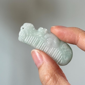 May include: A pale green jade comb pendant with carved details. The comb features a decorative design on the top and fine teeth. The pendant is held in a hand against a neutral background.