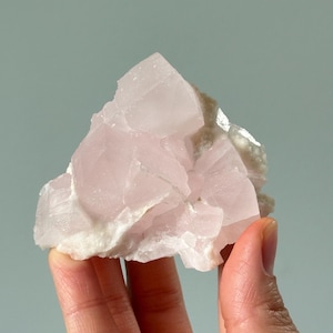 Peut inclure: Un amas de cristaux de quartz rose pâle à la surface rugueuse et texturée. Les cristaux sont posés sur un fond de matrice plus claire, blanc cassé. La forme générale est irrégulière, avec des bords tranchants et un aspect naturel et non poli.