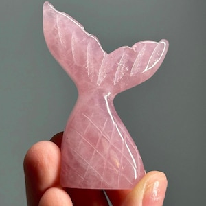 May include: A pink rose quartz mermaid tail sculpture. The tail is carved with detailed lines and has a smooth, polished finish. The sculpture is held in a hand against a gray background. The tail is approximately 3 inches tall.
