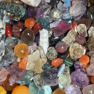 May include: A collection of various polished gemstones in shades of orange, purple, clear, and brown. The assortment includes spheres, heart-shaped stones, and carved figures. Some stones have visible inclusions and unique patterns.