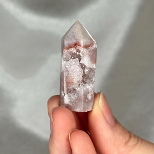 May include: A clear quartz crystal point with pink and white inclusions and red banding. The crystal is held between fingers against a soft gray background. The crystal is a natural, polished stone.