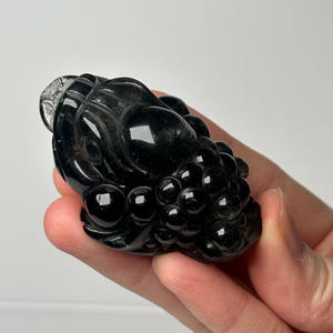 May include: A carved black crystal figurine of a mythical creature, possibly a dragon or a similar beast. The figurine is held in a hand, showcasing its intricate details and polished surface. The sculpture features a cluster of round shapes, possibly grapes.