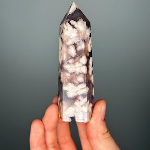 May include: A polished crystal tower with a pointed top. The crystal features a mix of white, brown, and gray patterns. The tower is held in a hand against a gray background. The crystal is approximately 5 inches tall.
