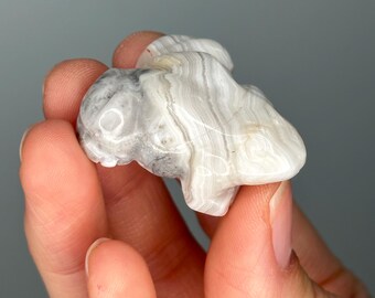 May include: A small, carved stone figurine of a bunny. The bunny is white and grey with a striped pattern. The figurine is held in a person's hand.