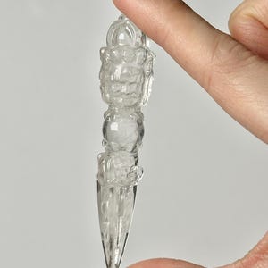 May include: A clear, intricately carved crystal object, held between fingers. The object has a rounded top with detailed carvings, a central sphere, and a pointed bottom. The crystal is transparent, allowing light to pass through, and appears to be a decorative item.