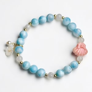 May include: A beaded bracelet featuring round, light blue marbled stones, small translucent white beads, and tiny gold-toned spacers. A carved pink shell-shaped charm and a small white flower-shaped charm with a pearl accent are attached.