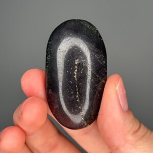 May include: A polished, oval-shaped stone with a dark, glossy surface. The stone features a prominent, light-colored, curved band that creates a visual contrast. Tiny gold flecks are scattered throughout the stone, adding a subtle sparkle.