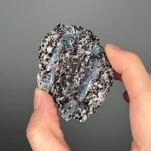 May include: A hand holds a rough, textured rock formation. The rock is primarily black and gray, with embedded blue, pink, and white crystalline structures. The background is a solid gray.