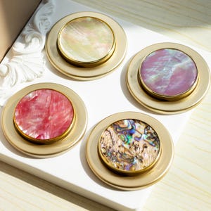 May include: Four round, gold-toned decorative knobs with iridescent mother-of-pearl inlays. The inlays feature different patterns and colors, including pink, white, and green.