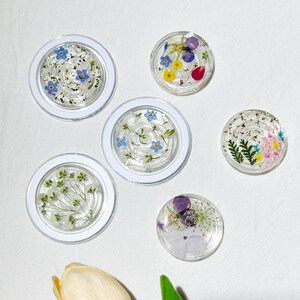 May include: A collection of clear resin coasters with dried flowers and gold flakes. The coasters feature various floral arrangements, including blue forget-me-nots, yellow and purple blooms, and delicate greenery, set within white circular frames.