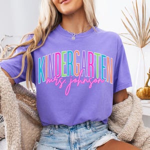 May include: A violet-colored t-shirt with the word "KINDERGARTEN" in a colorful, outlined font. Below the word is the name "Mrs. Johnson" in a cursive, pink font. The shirt is a short-sleeved tee.