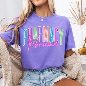 May include: A violet t-shirt with the words "PHARMACY Technician" in a colorful, layered design. The text is in shades of pink, green, and blue. The shirt is short-sleeved and made of a soft material.