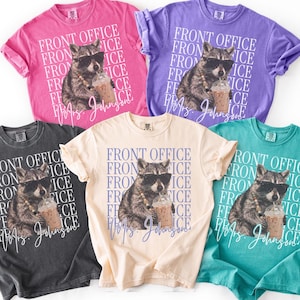 May include: Five short-sleeved t-shirts in pink, violet, dark gray, ivory, and teal. Each shirt features a graphic of a raccoon wearing sunglasses and holding an iced coffee, with the repeated text FRONT OFFICE and the script name Mrs. Johnson.