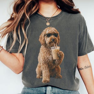 May include: Dark grey t-shirt featuring a brown dog wearing sunglasses and drinking an iced coffee. The dog graphic is centered on the shirt. The word "PEPPER" is printed on a light green rectangle at the bottom of the image.