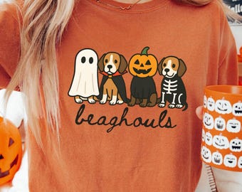 May include: Orange t-shirt with a Halloween-themed graphic of four dogs dressed as a ghost, vampire, pumpkin, and skeleton. The text "beaghools" is below the dogs. A matching orange mug with jack-o'-lantern faces is held in the hand.