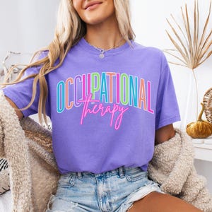 May include: A violet t-shirt with the words "OCCUPATIONAL therapy" in a colorful, outlined font. The shirt is paired with denim shorts. The image also includes a beige cardigan and a necklace.