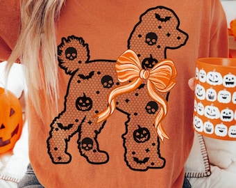 May include: Burnt orange t-shirt featuring a black lace poodle design with skull and pumpkin accents, and an orange bow. The shirt is part of a Halloween theme, with a matching pumpkin mug and jack-o'-lantern decor.