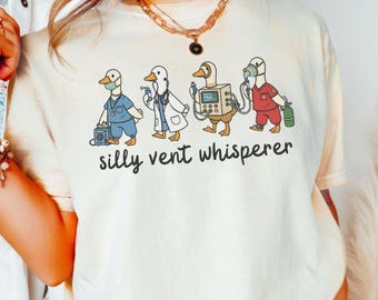 May include: Ivory-colored t-shirt featuring a whimsical illustration of four ducks dressed as medical professionals. The ducks are wearing scrubs, lab coats, and carrying medical equipment. The text "silly vent whisperer" is printed below the ducks.