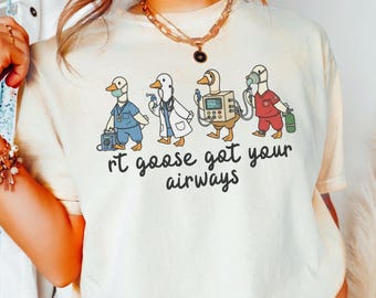 May include: Ivory-colored t-shirt featuring a cartoon design of four geese dressed as medical professionals. The geese are depicted with stethoscopes, medical equipment, and scrubs. The text "rt goose got your airways" is printed below the geese.