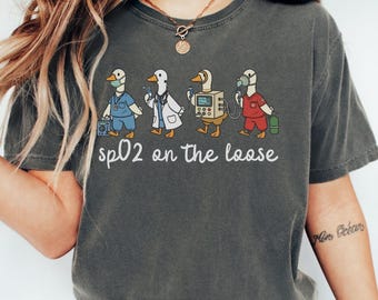 May include: Dark gray t-shirt featuring a whimsical illustration of four ducks dressed as medical professionals. The ducks are depicted in scrubs, lab coats, and with medical equipment. The text "sp02 on the loose" is printed below the ducks.