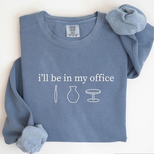 May include: Blue jean-colored sweatshirt with the text "i'll be in my office" and three white line-drawn images. The sweatshirt has a crew neck and long sleeves. A scrunchie in a matching blue color is on the right sleeve.