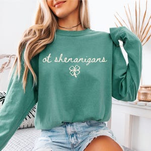May include: Light green sweatshirt with the text "at shenanigans" in a cursive font and a white four-leaf clover graphic. The sweatshirt is a crew neck style and has long sleeves. The bottom right corner of the image has the text "LIGHT GREEN".