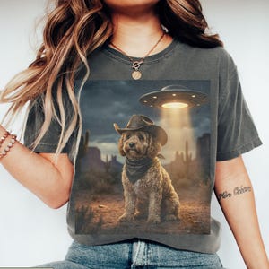 May include: Dark grey t-shirt featuring a graphic of a dog wearing a cowboy hat and bandana, with a UFO hovering above. The dog is set against a desert landscape with cacti and a cloudy sky. The word "PEPPER" is at the bottom.