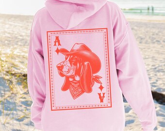 May include: Light pink hoodie with a large graphic on the back featuring a dog wearing a cowboy hat and bandana, framed within a playing card design. The text "LIGHT PINK" is at the bottom.