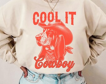 May include: Sand-colored sweatshirt with a red graphic of a dog wearing a cowboy hat and bandana. The text "COOL IT Cowboy" is above and below the dog illustration. The model is wearing denim shorts.