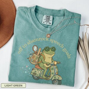May include: Light green t-shirt with the text "off to shamrock speech goals." The shirt features a cartoon frog riding a scooter, wearing shamrock sunglasses and carrying a bag labeled "SLP Life."