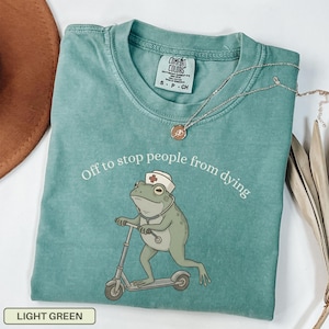 May include: Light green Comfort Colors t-shirt with the text "Off to stop people from dying." The shirt features a cartoon frog wearing a nurse's hat and riding a scooter. A silver necklace with a pendant is draped on the shirt.