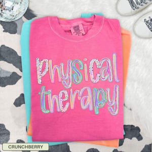 May include: A bright pink short-sleeve t-shirt with the text Physical therapy printed in a multi-colored, speckled, and outlined font across the chest. The shirt features a crew neckline and a Comfort Colors brand label on the inner collar.