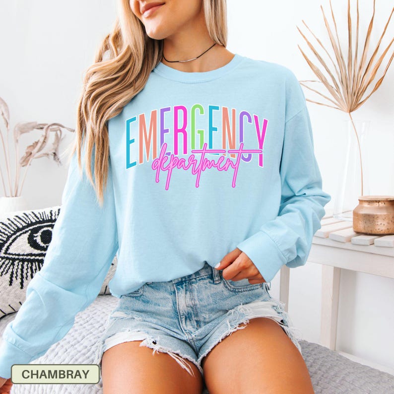 May include: Pale blue long-sleeved top featuring the text "EMERGENCY department" in a vibrant, multi-coloured design. The model is wearing denim shorts. The shirt is made of a soft, comfortable material.