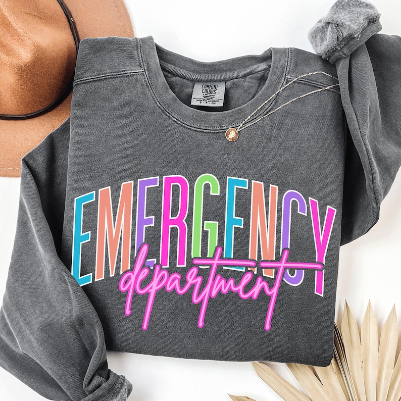 May include: A dark grey sweatshirt featuring the words "EMERGENCY department" in a vibrant, multi-coloured design. The text is outlined in bright colours, with a pink neon-style font for "department". A gold necklace and a brown hat are visible.