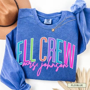 May include: A blue crewneck sweatshirt with the text "ELL CREW mrs. johnson" in colorful block letters and neon pink script. A gold necklace and a brown hat are visible in the background. The sweatshirt is labeled "FLO BLUE".