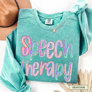 May include: A teal pigment-dyed crewneck sweatshirt featuring the text Speech Therapy in a multi-colored, marbled, and speckled font. The garment has a ribbed collar with a Comfort Colors brand label and long sleeves with ribbed cuffs.
