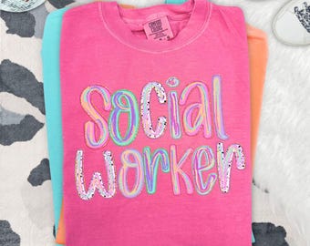 Puede incluir: Una camiseta de manga corta de color rosa brillante con el texto "SOCIAL WORKER" en una fuente multicolor, con remolinos y motas. La prenda tiene un cuello redondo y una etiqueta de la marca Comfort Colors impresa en el interior.