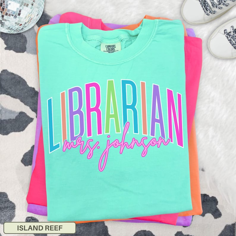 May include: A turquoise Comfort Colors t-shirt with the word "LIBRARIAN" in colorful block letters and "Mrs. Johnson" in cursive. The shirt is folded on top of other brightly colored shirts. The shirt's color is "Island Reef".