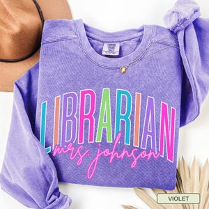May include: A violet-colored sweatshirt with the word "LIBRARIAN" in a colorful, outlined font. Below the word is the name "mrs. johnson" in a cursive font. A brown hat and gold necklace are visible in the background.