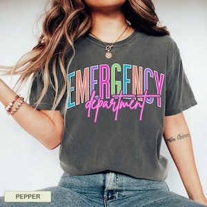 May include: A dark grey t-shirt featuring the words "EMERGENCY department" in a vibrant, neon-style font. The text is in pink, blue, green, and orange. The shirt is a casual style.
