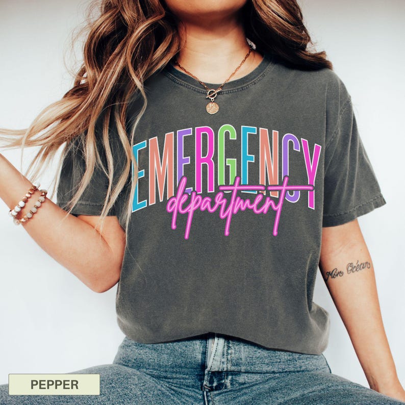May include: A dark grey t-shirt featuring the words "EMERGENCY department" in a vibrant, neon-style font. The text is in pink, blue, green, and orange. The shirt is a casual style.