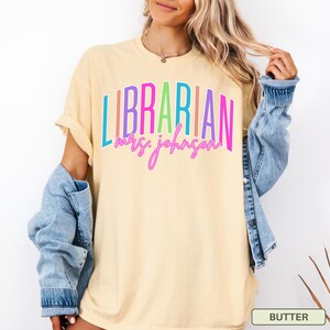 May include: A light yellow t-shirt with the word "LIBRARIAN" in colorful vertical stripes and the name "mrs. johnson" in a cursive font. The shirt is a casual style, with rolled-up sleeves.