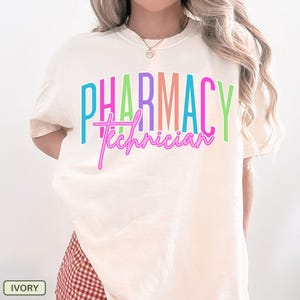 May include: Ivory-colored t-shirt with the words "PHARMACY Technician" in colorful, bold lettering. The text is in a mix of bright colors, with the word "PHARMACY" in a block style and "Technician" in a cursive font. The shirt is a casual style.