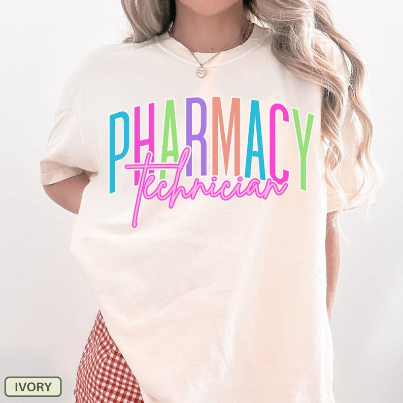 May include: Ivory-colored t-shirt with the words "PHARMACY Technician" in colorful, bold lettering. The text is in a mix of bright colors, with the word "PHARMACY" in a block style and "Technician" in a cursive font. The shirt is a casual style.