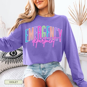 May include: A violet long-sleeved top featuring the text "EMERGENCY department" in a vibrant, neon-style font. The top has a crew neck and is a solid colour. The word "VIOLET" is displayed in a small box at the bottom.