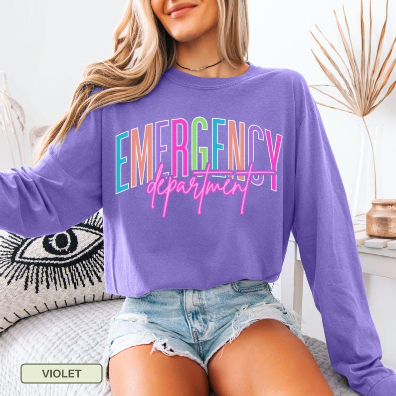 May include: A violet long-sleeved top featuring the text "EMERGENCY department" in a vibrant, neon-style font. The top has a crew neck and is a solid colour. The word "VIOLET" is displayed in a small box at the bottom.