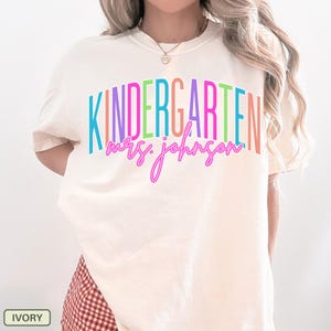 May include: Ivory-colored t-shirt with the word "KINDERGARTEN" in rainbow colors and "mrs. johnson" in a cursive font. The shirt is a comfortable, casual style, perfect for teachers.