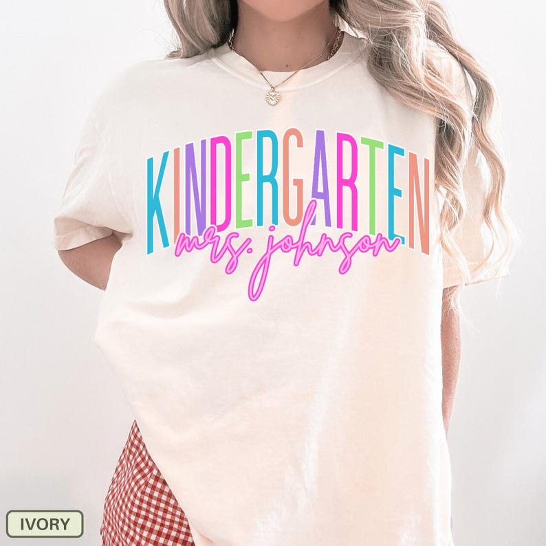 May include: Ivory-colored t-shirt with the word "KINDERGARTEN" in rainbow colors and "mrs. johnson" in a cursive font. The shirt is a comfortable, casual style, perfect for teachers.