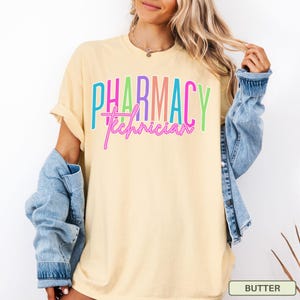 May include: A light yellow t-shirt with the words "PHARMACY Technician" in colorful block and script lettering. The shirt has short sleeves and a relaxed fit. A denim jacket is draped over the shoulders.