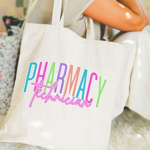 May include: Cream-colored tote bag with the words "PHARMACY Technician" in colorful, vertical text. The text is in shades of blue, green, pink, purple, and orange. The bag has two handles and a small tag that says "TOTE BAG".